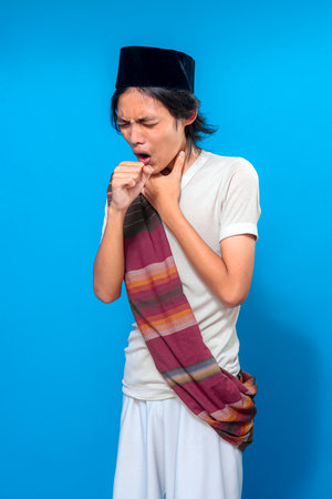 Traditional Indonesian man wearing black peci and colorful sarong coughs and holds his throat, expressing discomfort or sore throat with a painful, emotional facial expression.の写真素材