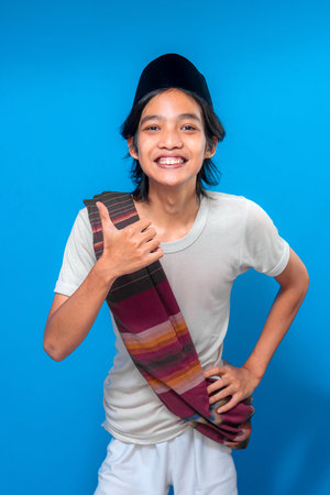 A cheerful Indonesian young man wears a traditional peci and loosely draped sarong while smiling widely and giving a thumbs-up, expressing positivity and friendliness with full energy.の写真素材