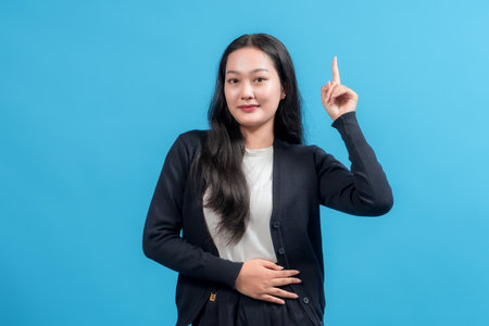 Young woman pointing upward with one hand, posing confidently against bright blue background, symbolizing idea, innovation, or emphasis. Perfect for education, communication, and marketing themes.の写真素材