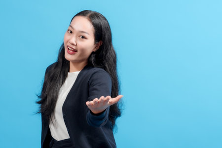 A cheerful young woman in a dark blue sweater and a white shirt gestures warmly while standing against a vivid blue background. The composition exudes a friendly and inviting atmosphere.の写真素材