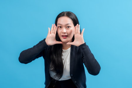A young woman with long black hair is cupping her hands around her mouth, as if shouting or calling out, against a bright blue background, conveying enthusiasm and energy.の写真素材