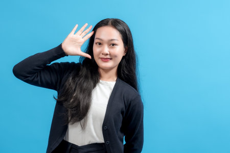 Portrait of a cheerful young woman with long black hair waving hand in greeting against a blue background, showcasing positive emotions, friendliness, and modern fashion styling.の写真素材