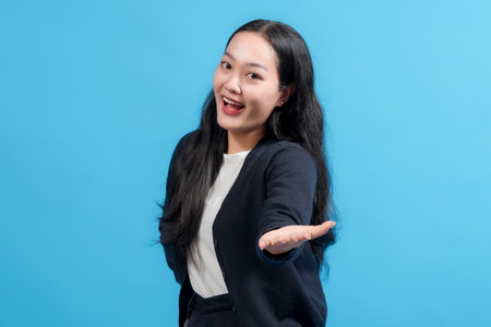 A cheerful young woman in a dark blue sweater and a white shirt gestures warmly while standing against a vivid blue background. The composition exudes a friendly and inviting atmosphere.の写真素材