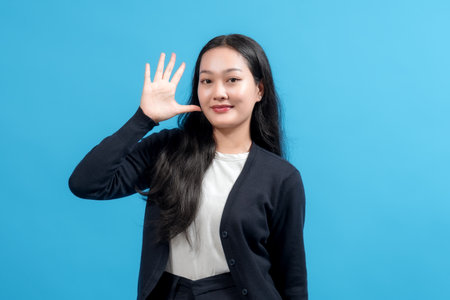 Portrait of a cheerful young woman with long black hair waving hand in greeting against a blue background, showcasing positive emotions, friendliness, and modern fashion styling.の写真素材