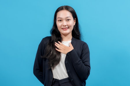 Young woman with a sincere expression placing hands on her chest, standing confidently against a solid blue background, conveying gratitude, appreciation, or heartfelt emotion.の写真素材