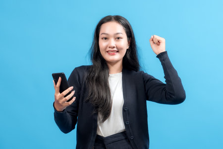 Happy young woman celebrating success while holding smartphone, raising fist in victory gesture, standing against blue background. Concept of good news, achievement, and online success.の写真素材
