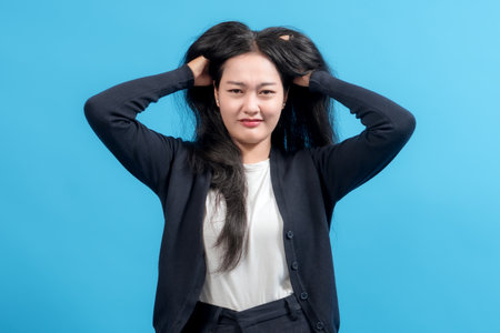 Frustrated woman in casual clothing pulling hair with stressed expression, standing against blue background. Concept of pressure, anger, mental breakdown, and work-life struggles.の写真素材