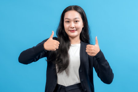 A confident young woman with long hair smiling and showing a thumbs-up gesture, expressing positivity and encouragement on a blue background.の写真素材