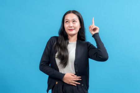 Young woman pointing upward with one hand, posing confidently against bright blue background, symbolizing idea, innovation, or emphasis. Perfect for education, communication, and marketing themes.の写真素材