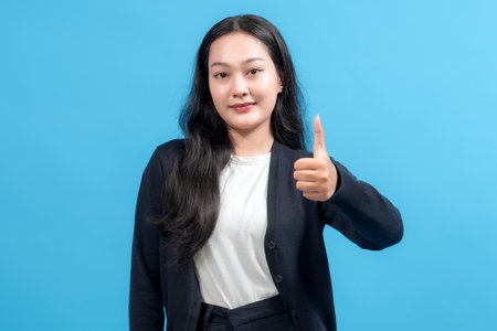 Smiling woman giving thumbs up gesture against blue background, showing approval, positivity, and success. Ideal for concepts of agreement, encouragement, or customer satisfaction.の写真素材