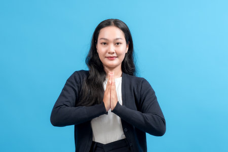 Professional woman dressed in business attire expressing gratitude with a confident smile. Set on plain blue background, the image captures positivity, respect, and a professional demeanor.の写真素材