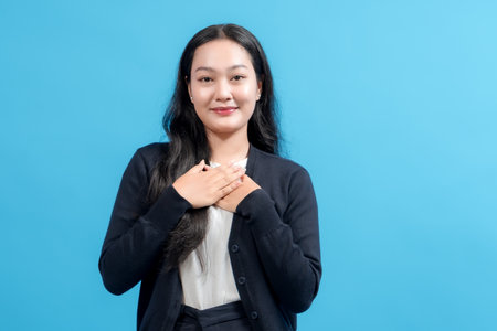 Young woman with a sincere expression placing hands on her chest, standing confidently against a solid blue background, conveying gratitude, appreciation, or heartfelt emotion.の写真素材