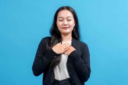 Woman with eyes closed and hands on chest, showing gratitude and peace against blue background. Perfect for concepts of thankfulness, mindfulness, and emotional well-being.の写真素材