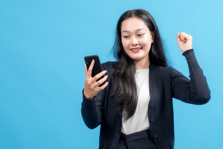 Happy young woman celebrating success while looking at smartphone, raising fist in victory gesture, standing against blue background. Concept of good news, achievement, and online success.の写真素材