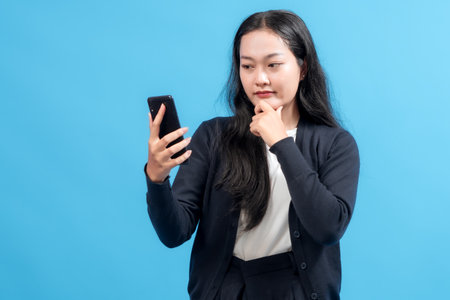 Confident businesswoman holding smartphone, thinking with hand on chin, standing against blue background. Represents decision-making, communication, and technology in business.の写真素材