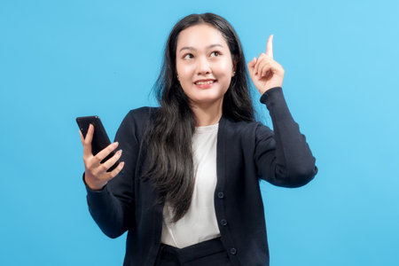 Smiling businesswoman holding smartphone, pointing up with finger, standing against blue background. Symbolizes idea, innovation, communication, and modern technology in business.の写真素材