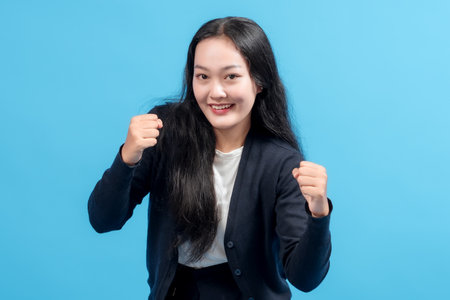 Happy young Asian woman celebrating success with raised fists, wearing casual outfit, standing against blue background. Concept of achievement, victory, motivation, and positive energy.の写真素材