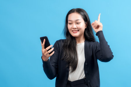 Smiling businesswoman holding smartphone, pointing up with finger, standing against blue background. Symbolizes idea, innovation, communication, and modern technology in business.の写真素材