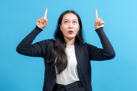 Surprised woman pointing up with both index fingers, looking upward against a blue background. Perfect for concept ideas, promotions, and advertising designs.の写真素材