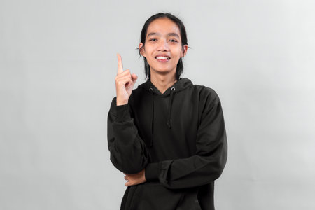 Young Asian man in black hoodie smiling and pointing upward with index finger, looking confident and expressive, symbolizing idea, inspiration, or smart solution, on gray background.の写真素材