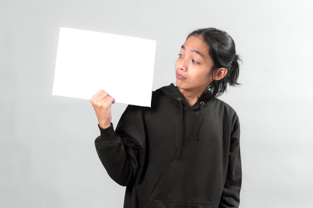 Asian man in black hoodie holding a blank white sign while looking at it with a curious expression, isolated on gray background, perfect for advertisement or message mockup.の写真素材