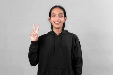 Portrait of young Asian man in black hoodie smiling and showing peace sign with two fingers. Standing against gray background, expressing positivity, happiness, and friendly gesture.の写真素材