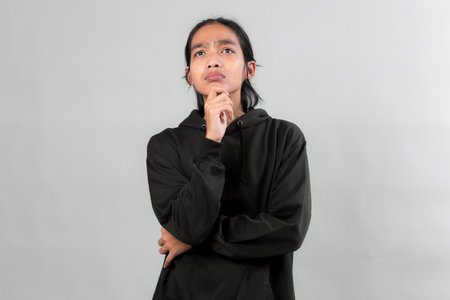 Young Asian man in black hoodie looking up with thoughtful expression, hand on chin, isolated on gray background, concept of decision making or ideas.の写真素材