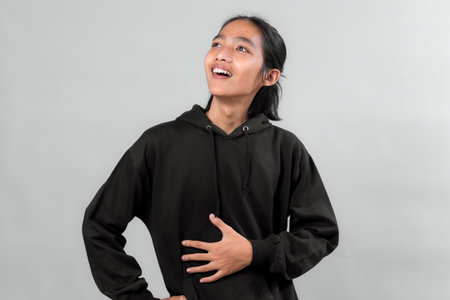 Young Asian person in a black hoodie holding stomach with one hand and looking up with joyful surprise, showing excitement or amazement, isolated on a neutral gray background.の写真素材