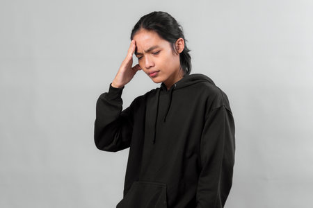 Asian man in black hoodie looking stressed with hand on forehead, eyes closed, and furrowed brows. Studio portrait on gray background expressing headache, tension, or anxiety.の写真素材