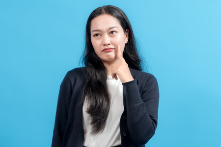 A young woman with long hair and a thoughtful expression, standing confidently against a solid blue background, showcasing a casual yet stylish appearance while making a hand gesture.の写真素材
