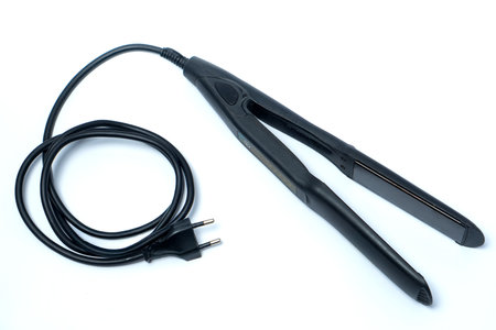 A black electric hair straightener with a long cord and plug placed on a white background,...
