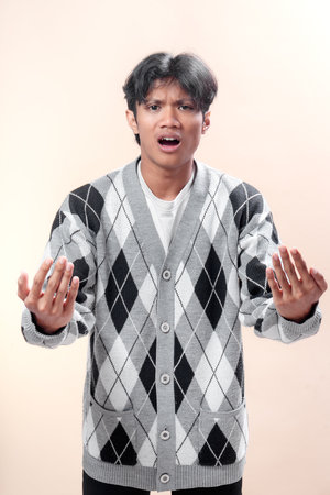 Asian man in gray argyle cardigan with open mouth and raised hands, showing confused and frustrated...
