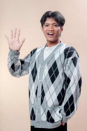 Asian man in gray argyle sweater smiling while raising five fingers with one hand, showing cheerful gesture, positive attitude, confidence, youthful spirit, and friendly professional image.の写真素材