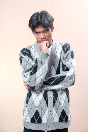 An Asian man in a gray argyle cardigan poses with his hand on his chin and arms crossed, showing a serious thoughtful expression, focus, confidence, and determination.の写真素材