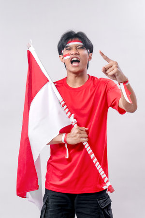 A young Indonesian man passionately celebrates Independence Day, holding the flag while shouting and pointing in excitement. Dressed in a red shirt, with face paint and wristbands, he shows his patriotic spirit.の写真素材