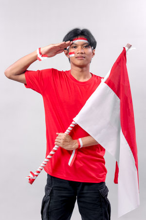 A young Indonesian man salutes proudly while holding the Indonesian flag, embodying patriotism and respect for the nation. His red shirt, face paint, and wristbands symbolize national pride.の写真素材