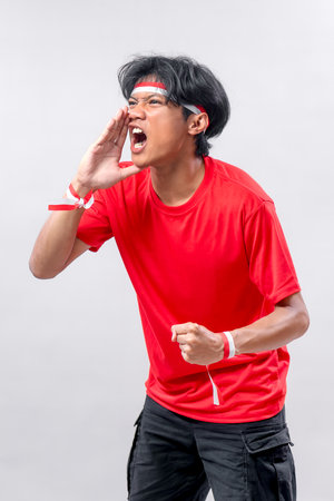 Young Indonesian man shouting with clenched fist, wearing red t-shirt and red-white accessories. Expressing patriotic spirit during Independence Day celebration.の写真素材