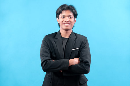 A confident young man in a black suit smiles with folded arms against a blue background,...