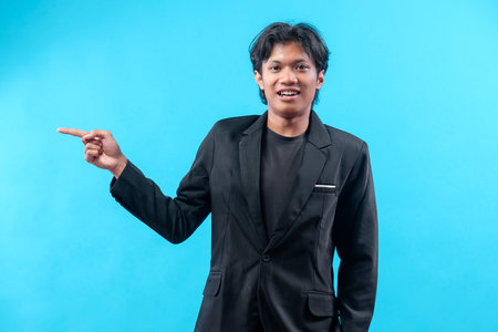 A confident young man wearing a black suit and t-shirt is pointing to the side while smiling, expressing positivity and professionalism against a plain blue background.の写真素材