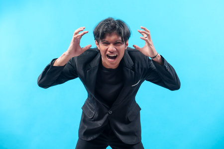 An Asian man in a black suit shows an angry expression with raised hands and open mouth, symbolizing stress, frustration, and emotional tension in a professional concept.の写真素材