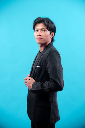 A young man in a black suit posing with hands on jacket, looking confident and serious, symbolizing ambition, professionalism, and modern business style on blue background.の写真素材