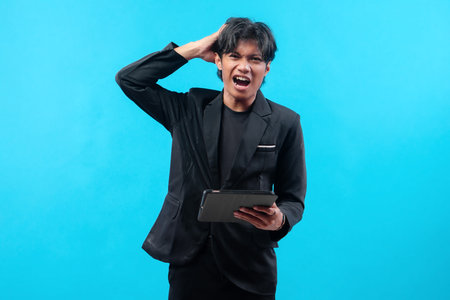 A young man in a black suit holding a tablet and scratching his head with an angry expression.の写真素材