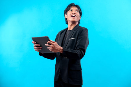 A cheerful young Asian man in a black suit holds a digital tablet and looks up with a bright smile, showing a blend of positivity, confidence, and professional enthusiasm.の写真素材