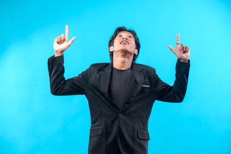 A young man in a black suit and t-shirt points both fingers upward while looking above, showing...