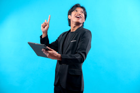 A happy young Asian man in a black suit smiles brightly while pointing his index finger upward and holding a digital tablet, showing inspiration, confidence, and bright ideas.の写真素材