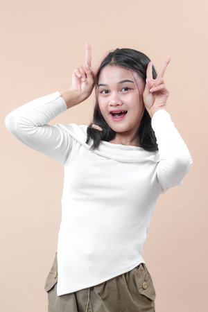 An Asian woman in a casual white shirt smiling with open mouth while raising both hands making playful peace sign gestures against a plain background.の写真素材