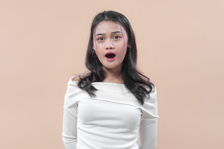 Asian woman with long black hair wearing a white off shoulder top, looking surprised with open mouth expression against plain background.の写真素材