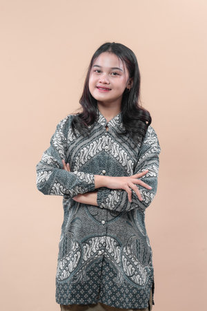 A confident young Asian woman wearing traditional batik dress stands with folded arms, smiling...