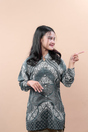 An Asian woman wearing a patterned batik shirt points to the side with a friendly smile, showing elegance, confidence, and positive body language in a modern professional concept.の写真素材