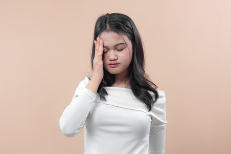 Asian woman with long black hair in a white off shoulder top, touching her face with closed eyes, showing tired and stressed expression against plain background.の写真素材